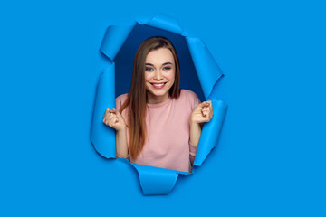 Portrait of beautiful happy smiling caucasian woman raises clenched fists and poses in blue paper hole.