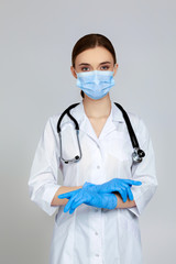 Portrait of beautiful woman doctor in white lab coat, mask and rubber gloves and stethoscope isolated on gray background