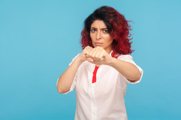 Confident hipster woman with fancy red hair in white shirt clenching fists to camera, courageously fighting threat, expressing confidence and strength. indoor studio shot isolated on blue background