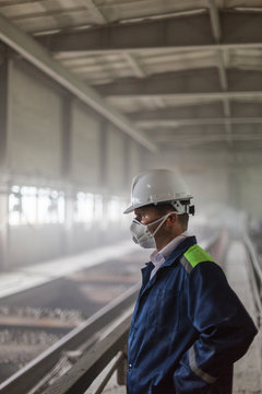 Mining Engineer In Yellow-blue Uniform And White Helmet And Respirator Inspects Dusty And Dirty Mining Workshop