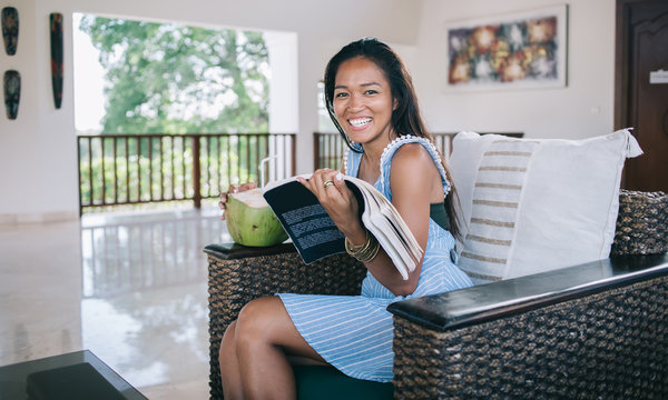 Long haired Asian woman with book and coconut cocktail smiling at camera