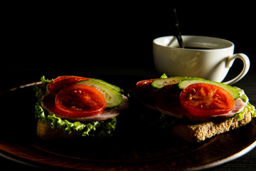 delicious sandwiches with ham, lettuce, cucumber, tomato and a Cup of coffee on a dark background.