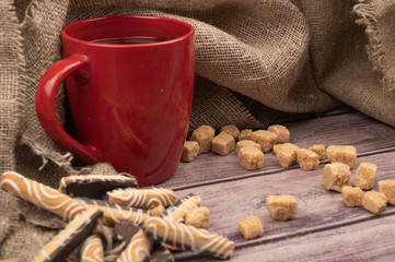 Red ceramic mug with tea, cookie sticks with chocolate and pieces of brown cane sugar on the background of a cloth with a rough texture. Close up.