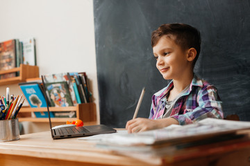 cute little boy schoolboy sitting at a table opposite a black board and studying online from his apartment
