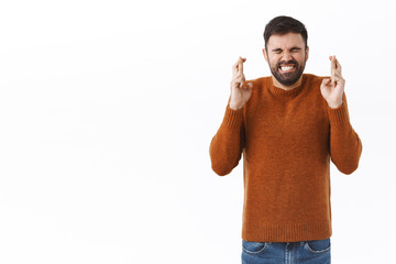 Fototapeta premium Portrait of excited, handsome bearded man wishing with all his heart for dream come true, close eyes and clench teeth put effort into praying, cross fingers good luck hopeful, white background