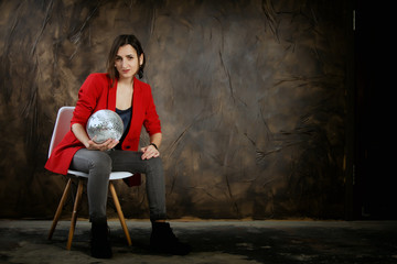 Attractive woman in a red jacket sitting against a dark wall. Young mysterious girl with glass ball in hands, fortune telling Dark tones. Copy space