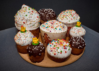 Sweet Easter cakes in white protein and chocolate glaze, decorated with pastry topping and yellow chicken on a round wooden plate on a gray background