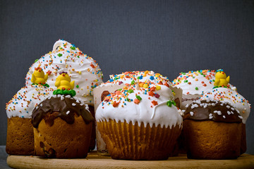 Sweet Easter cakes in white protein and chocolate glaze, decorated with pastry topping and yellow chicken on a round wooden plate on a gray background