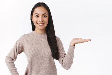 Cheerful asian woman in casual sweater, introduce product or banner, present corporate advertisement, smiling joyfully and polite, holding palm right over white background