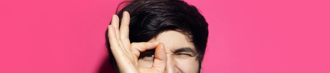 Close-up of young guy showing OK gesture on eyes, panoramic portrait on pink background.
