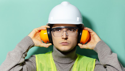 Portrait of young construction engineer worker wearing safety helmet, goggles, jacket and headphones on background of aqua menthe color.