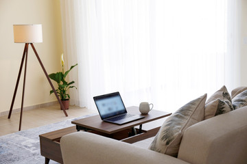 Home office concept. Designated work from home area in living room. Modern laptop and cup of hot beverage on wooden table. Close up, copy space, background.