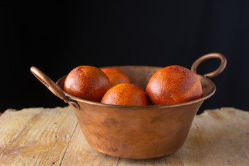 Close-up of copper bowl with blood oranges on rustic wooden table, with black background, horizontal