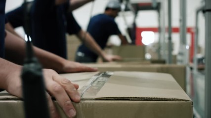 An operator closes a cardboard box with adhesive tape in a warehouse, after filled it with products. The operator puts tape on the cardboard box in the packaging line.Automation shipping logistics - Powered by Adobe