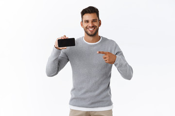 Cheerful smiling cute man give advice check-out good promo on mobile screen. Handsome male model in grey sweater holding smartphone horizontally pointing telephone screen satisfied