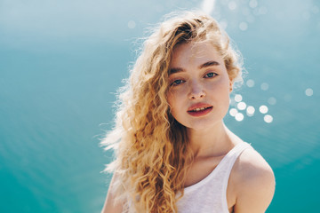 Mysterious curly blonde on a background of blue sea looks at the camera.