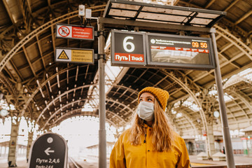 Young woman in protective medical mask dressed in yellow windbreaker and knitted cap at the empty railway station waiting for a train to drop off the city because of coronavirus pandemic. Covid-19 