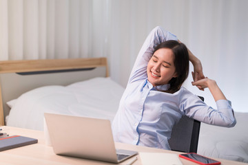Attractive Asian woman works from home with laptop on desk in bedroom. Young girl raises arms, stretching to relieve pain from office syndrome. Shelter in place from COVID-19 and Stay Home concept.