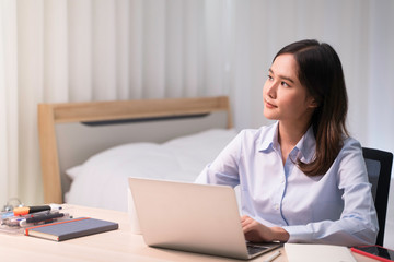 Attractive Asian woman works from home with laptop on desk in bedroom. Young girl smiling with happy face, looking outside and thinking about work. Shelter in place from COVID-19 and Stay Home concept