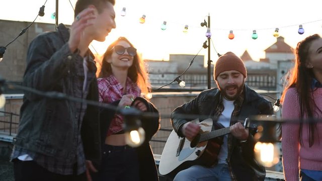 Holidays on the rooftop at sunset. A cheerful group of friends is resting during party with a guitar and snacks.