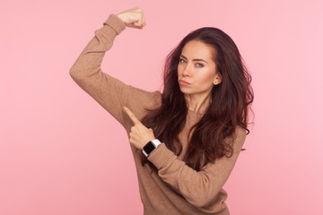 Concept of woman power and independence. Determined brave young lady with brunette hair pointing at biceps, expressing strength and confidence, feminist views. studio shot isolated on pink background