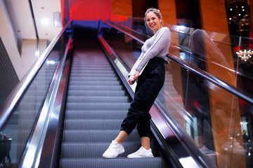 Obraz premium Young woman rises on an escalator to the mall and holds a laptop in her hands. career advancement concept. Woman listening to music using her wireless earphones.