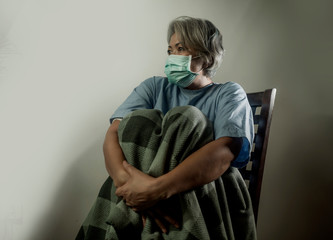 covid-19 virus spreading disease - scared and sad mature woman with grey hair in hospital patient gown and mask infected by coronavirus feeling sick and in panic