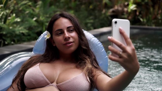 A beautiful woman is relaxing in the pool at a luxury beach resort spa hotel taking a selfie on her phone