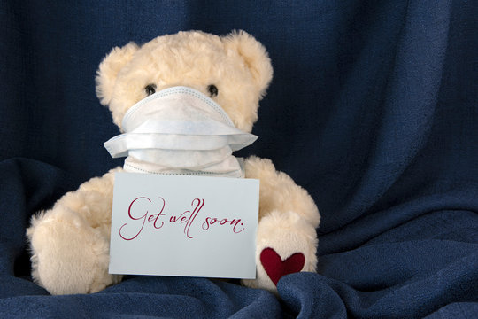 Teddy bear with facemask, blue cloth background, words GET WELL SOON on card