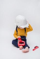 The kid plays a builder. Baby boy sits in a helmet with a shurovert. Baby boy on a white background in a yellow bodysuit, blue pants and a white hard helmet