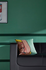 The interior photo of a black couch with a colorful pillow and a modern picture on the green wall. The pillow-case is decorated with a comics painting of a shaving lady.