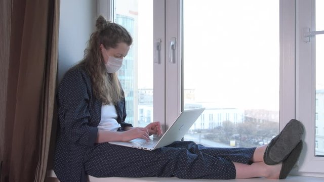 Bored woman wearing mask looking at window and working on laptop from home due to coronavirus self isolation. Sitting on window sill. Authentic home workplace. Handheld shot. Coronavirus outbreak.