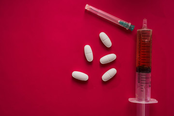 large and small pills scattered on a red background and a syringe top view