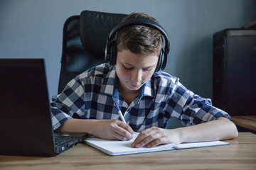 A teenager in a checked shirt and headphones is studying at home on a laptop. He sits at his Desk and makes notes in a notebooklaptop. Topic of distance learning