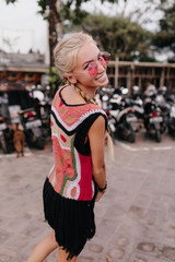 Fototapeta na wymiar Pretty girl playfully looking over shoulder with smile while spending time on the street. Outdoor shot of slim lady with braids wears pink sunglasses having fun in weekend.