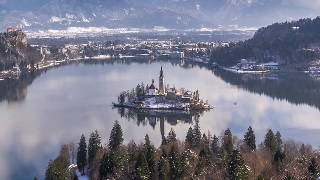 Time lapse zoom in view of Lake Bled island church and boats in winter landscape. Calm lake water reflects sky clouds above, snow covered mountains in the background