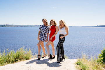 Shoot of three beautiful girls outdoors by the river. Female friends relaxing by the river and smiling. Girlfriends are stand on the pier, joy, fun, hands up. Girls with drinks in dresses. Summer