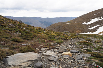 The mountain landscape with the light grey rocks, the slope covered by low grass and white snow on the cloudy day.