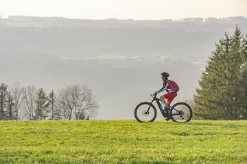 Naklejka premium pretty senior woman riding her electric mountain bike in early springtime in the Allgau mountains near Oberstaufen, in warm evening light below the spectacular snow capped mountains of Nagelfluh chain