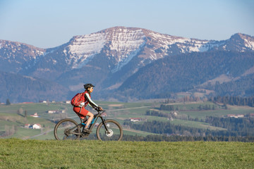 Obraz premium pretty senior woman riding her electric mountain bike in early springtime in the Allgau mountains near Oberstaufen, in warm evening light below the spectacular snow capped mountains of Nagelfluh chain