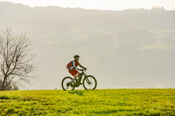 Fototapeta na wymiar pretty senior woman riding her electric mountain bike in early springtime in the Allgau mountains near Oberstaufen, in warm evening light below the spectacular snow capped mountains of Nagelfluh chain