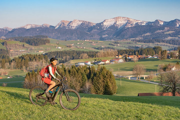 pretty senior woman riding her electric mountain bike in early springtime in the Allgau mountains near Oberstaufen, in warm evening light below the spectacular snow capped mountains of Nagelfluh chain