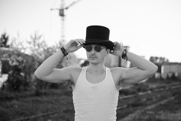 Three-quarter portrait of young man, wearing white top with sunglasses, holding his black classic hat, thinking. Black and white picture of creative man on abandoned construction site area. Art-house.