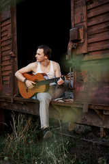 Young handsome man, wearing white t-shirt and grey pants, sitting inside old railway carriage, playing guitar, composing music Brunette male musician singer with guitar on abandoned train in summer.