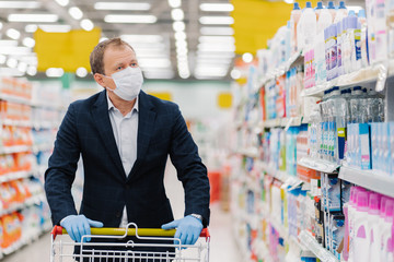 Serious attentive man chooses household detergents in store, walks with shopping cart, buys...