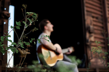 Young handsome man, wearing white t-shirt and grey pants, sitting inside old railway carriage, playing guitar, composing music Brunette male musician singer with guitar on abandoned train in summer.