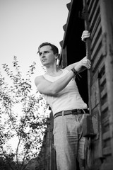 Three-quarter portrait of young man, wearing grey pants, white top, standing inside old railway carriage, holding to doors. Black and white picture of creative man on abandoned train area. Art-house.