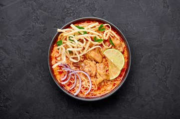 Khao Soi Gai or Thai Coconut Curry Noodle Soup in black bowl at dark slate background. Khao Soy is Thailand cuisine dish with noodles, chicken meat, red chillies paste and spices. Copy space