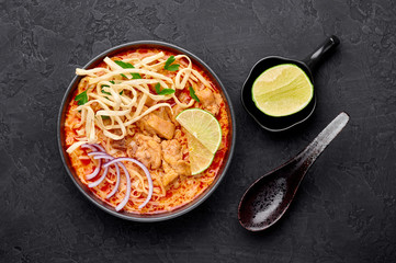 Khao Soi Gai or Thai Coconut Curry Noodle Soup in black bowl at dark slate background. Khao Soy is Thailand cuisine dish with noodles, chicken meat, red chillies paste and spices.