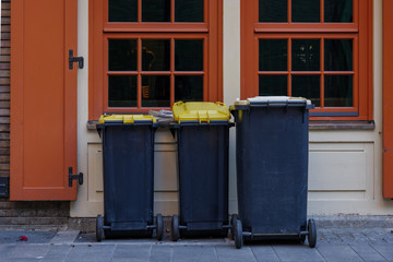 Naklejka premium Three black waste garbage bins on sidewalk in front of windows of buildings.